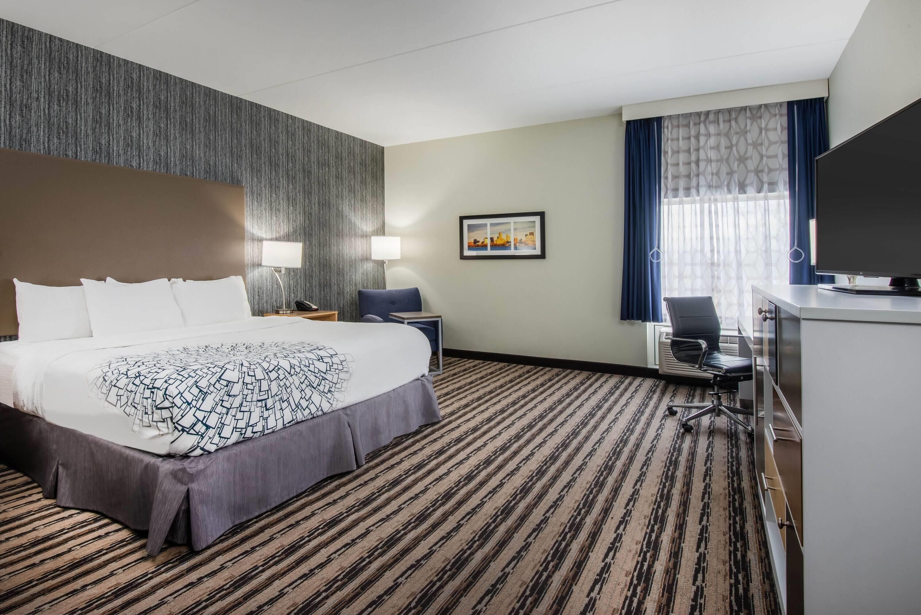 Une chambre d’hôtel avec un grand lit, un bureau avec une chaise, une télévision et une fenêtre avec des rideaux.
