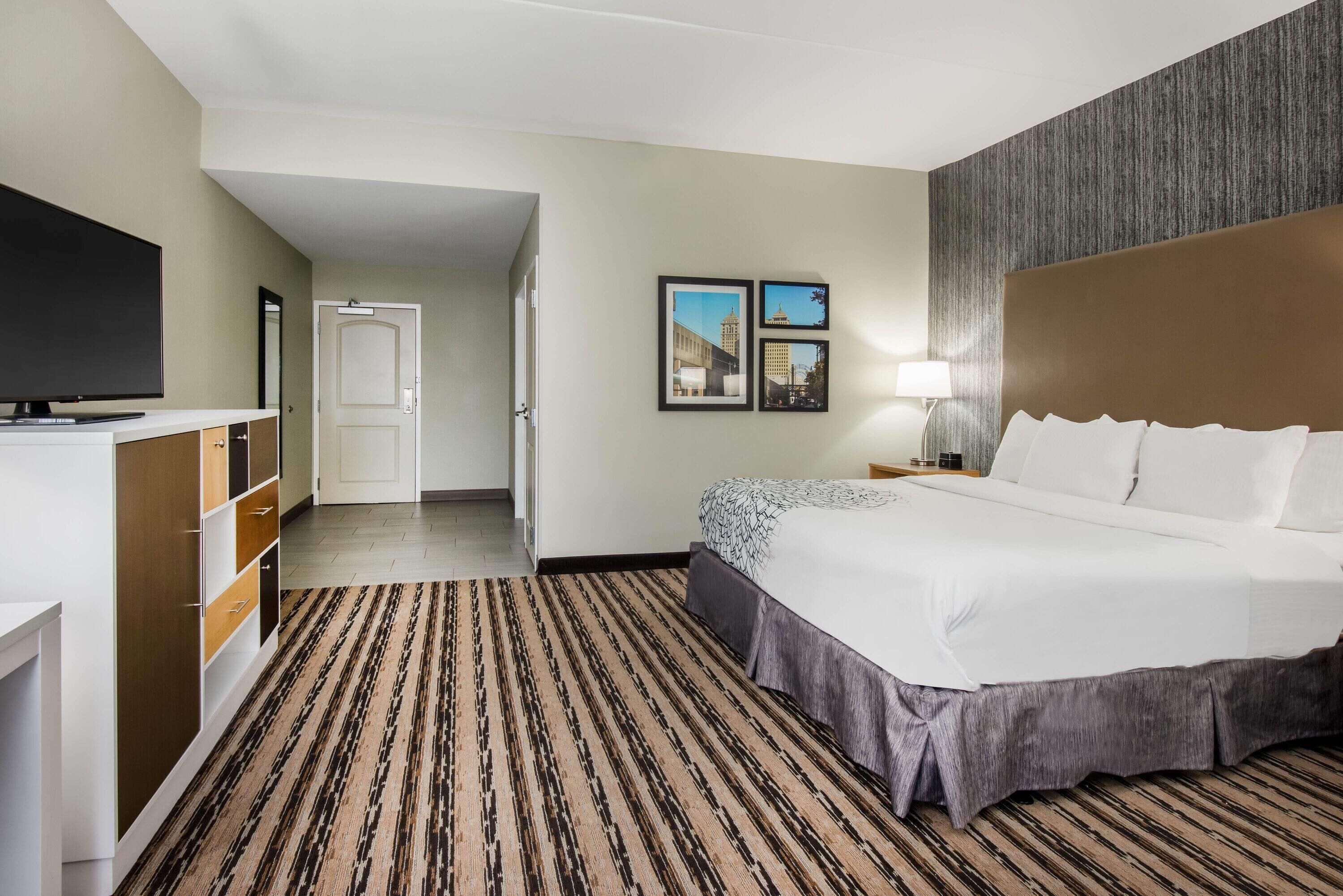 A hotel room with a large bed, a TV, a desk, and a wardrobe. There are framed pictures on the wall and a lamp on the bedside table.