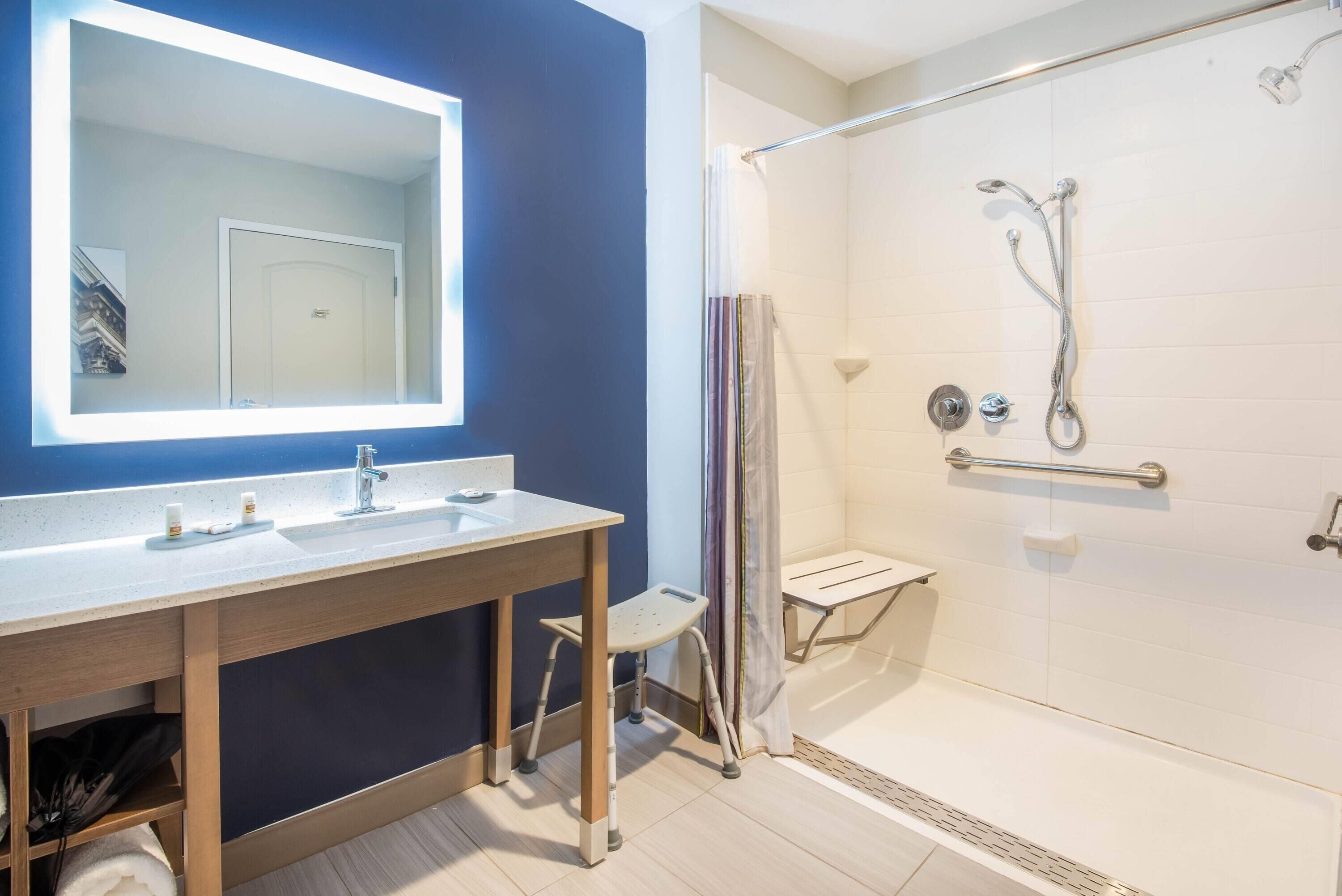 A bathroom with a large mirror, a sink, a shower with a glass door, and a small stool.