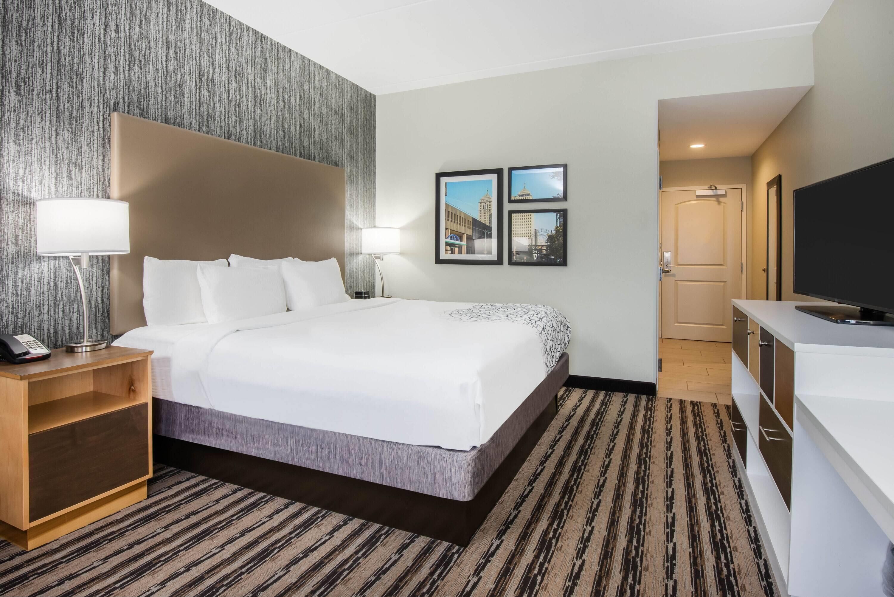 Una habitación de hotel con una cama grande, una mesita de noche con lámpara, televisión y un mueble pequeño.