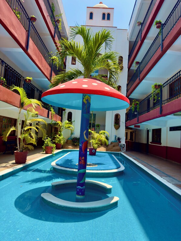 A swimming pool with a colorful umbrella, surrounded by potted plants and a building with balconies.