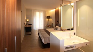 A modern hotel room with a large bed, a bedside table, a desk, a chair, a large mirror, and a bathroom with a sink and mirror. - Hotel Raval de la Mar (Vila-Seca)