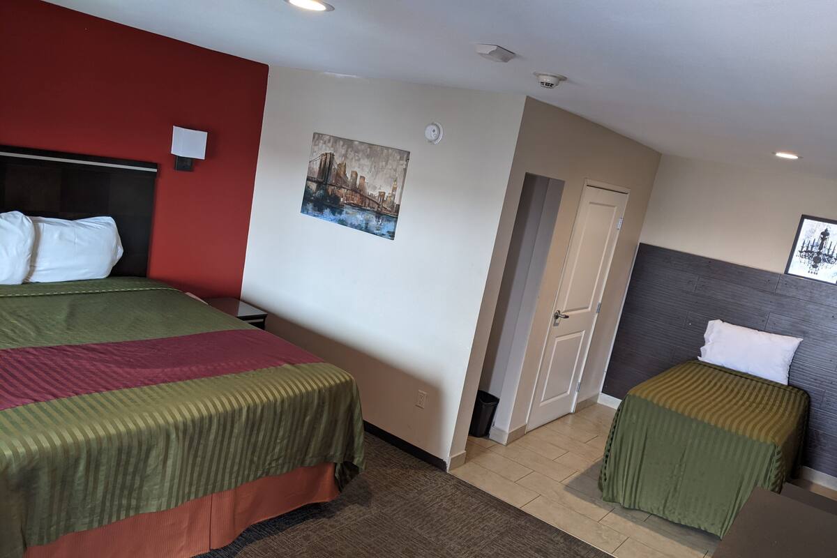 Une chambre d’hôtel avec deux lits, un mur d’accent rouge, un tableau représentant un paysage urbain et une porte donnant sur une autre pièce.