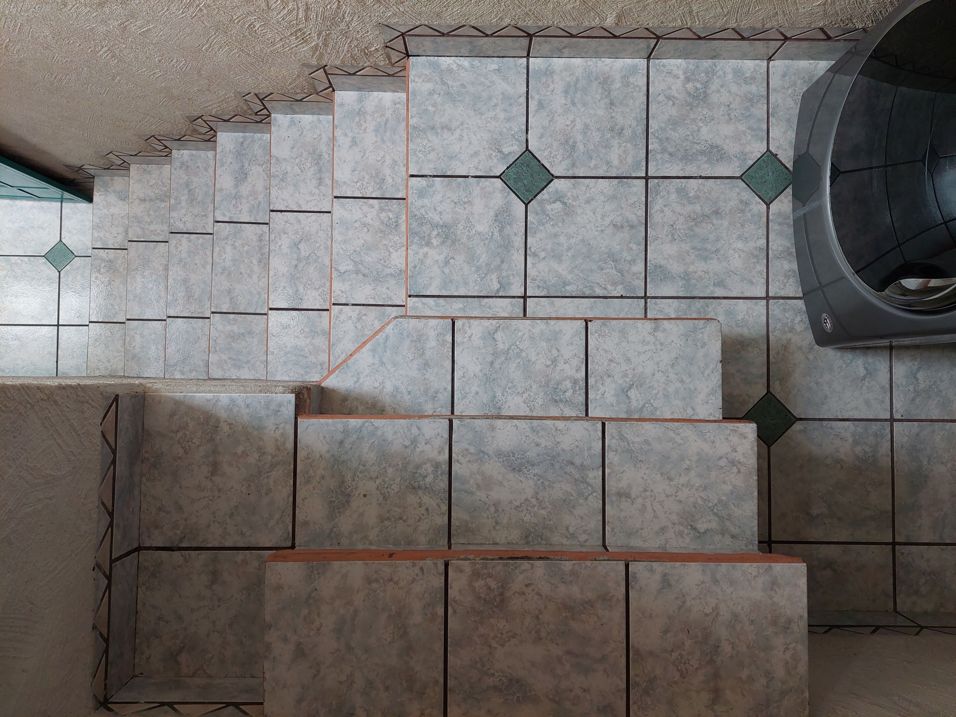 Un escalier orné d’un motif géométrique en carreaux, menant à une pièce où se trouve un grand appareil électroménager de couleur sombre.