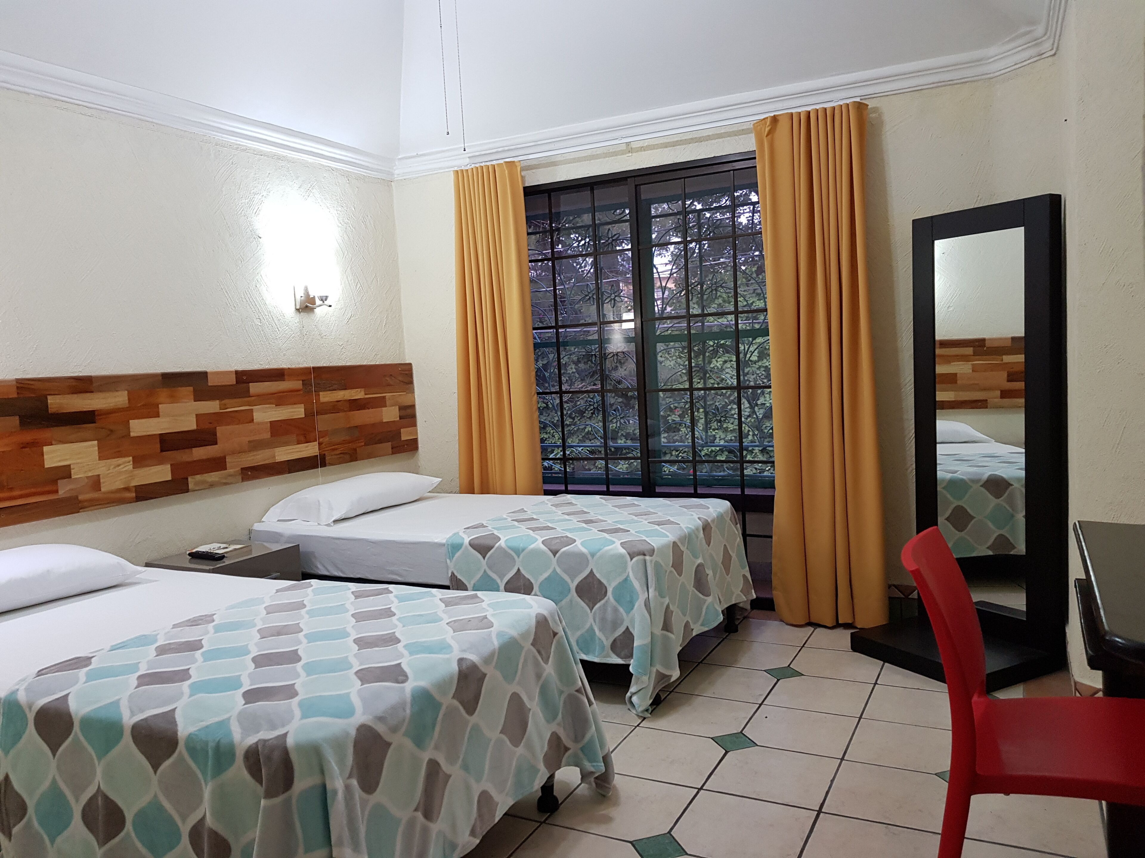 A hotel room with two beds, a wooden headboard, a window with curtains, a mirror, and a red chair.
