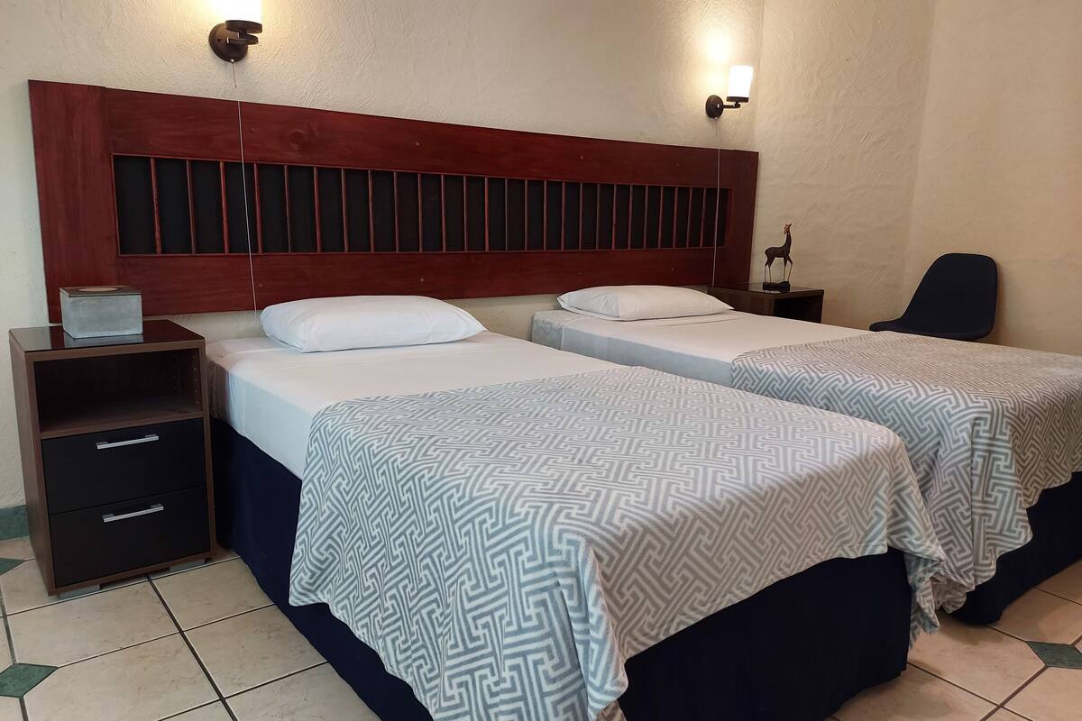 A hotel room with two beds, a nightstand, a chair, and a wall-mounted light fixture.