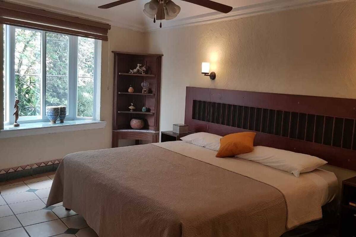 A neatly made bed with a headboard, two bedside tables, a nightstand, a window with blinds, and a shelf with decorative items.