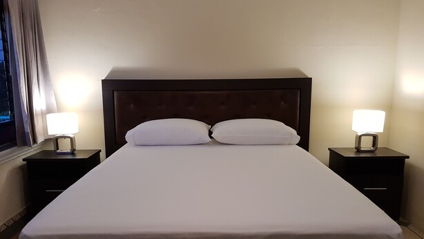 A neatly made bed with white linens, two bedside tables with lamps, and a window with curtains. - Aurora Express (Guatemala City)