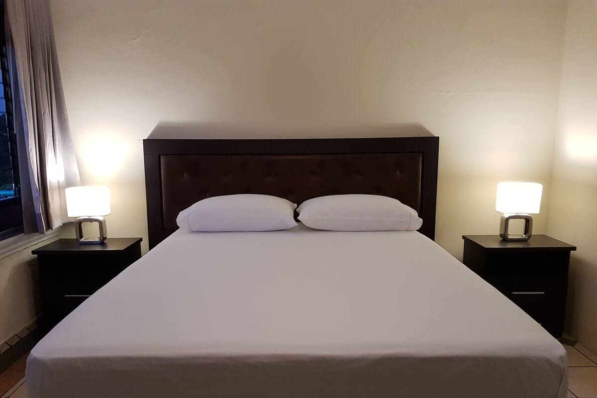 A neatly made bed with white linens, two bedside tables with lamps, and a window with curtains.