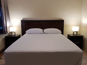 A neatly made bed with white linens, two bedside tables with lamps, and a window with curtains. - Aurora Express (Guatemala City)