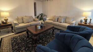 A living room with two sofas, a coffee table, and a blue armchair. There are lamps on side tables, a chandelier, and a patterned rug. - Aurora Express (Guatemala City)