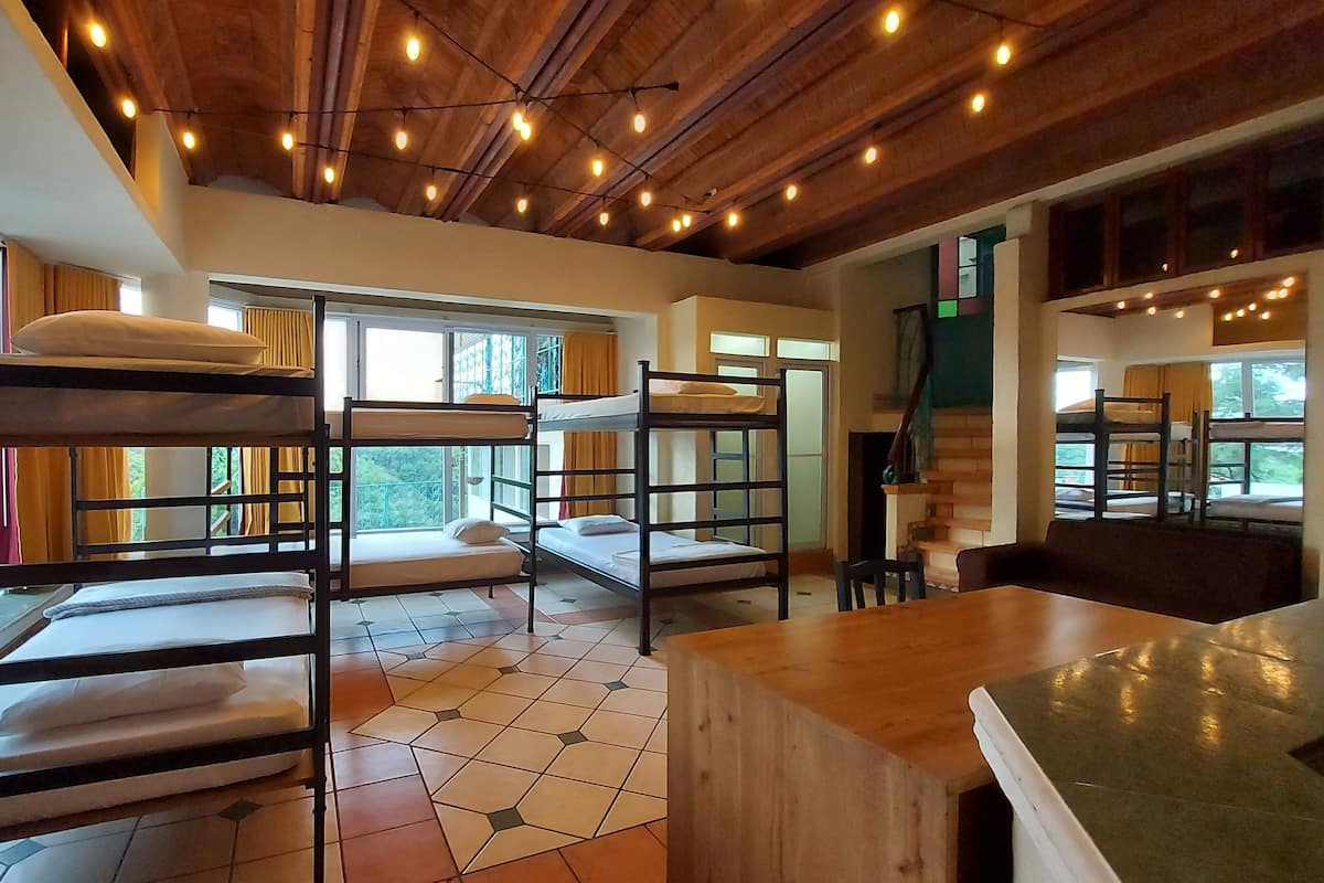 A dormitory-style room with bunk beds, large windows, and a wooden ceiling with exposed beams.