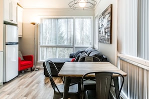 A compact kitchen with a refrigerator, a dining table with chairs, a red sofa, and a large window with blinds. - Le Villageois by Gestion ELITE  (Mont-Tremblant)