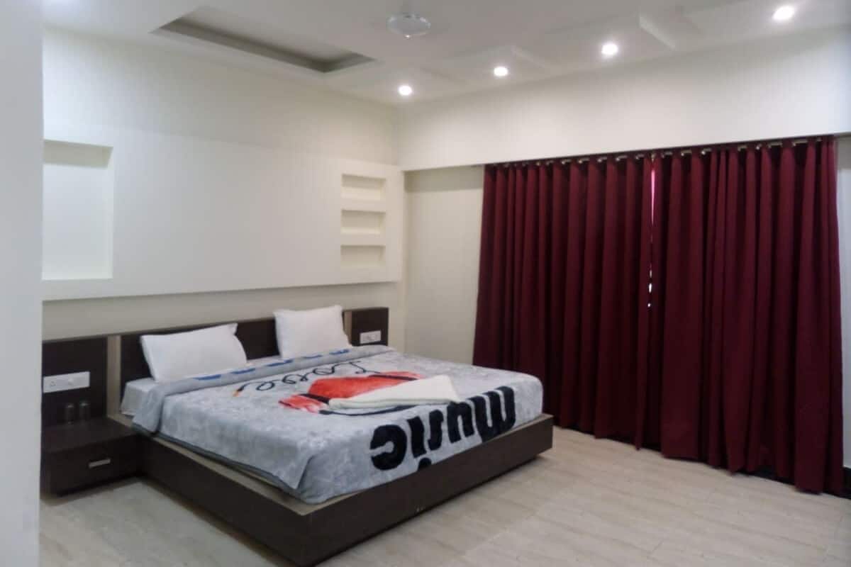 Un dormitorio con cama, mesita de noche, estante en la pared y ventana con cortinas rojas.