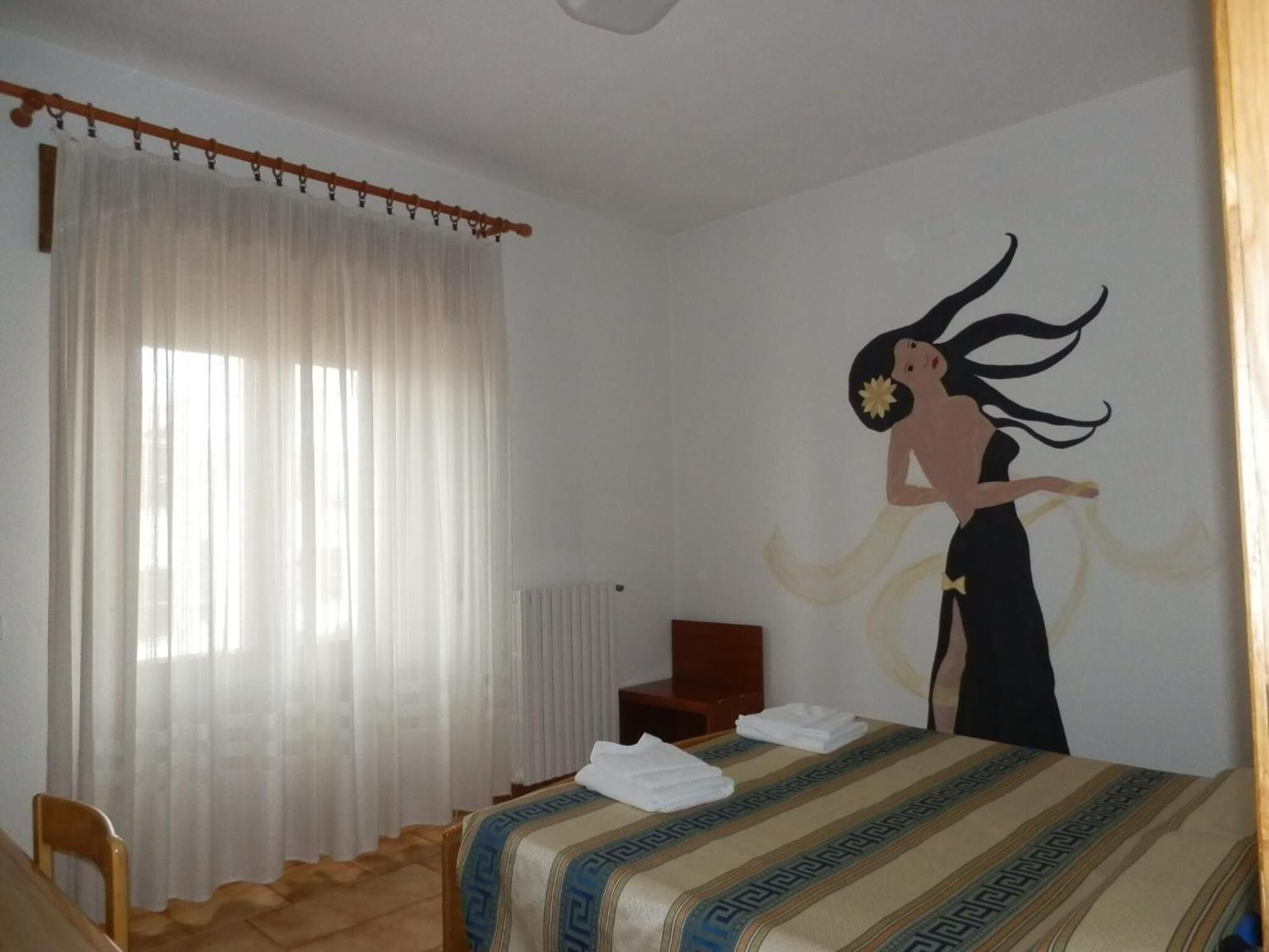 A hotel room with a bed, a chair, a radiator, and a wall with a mural.
