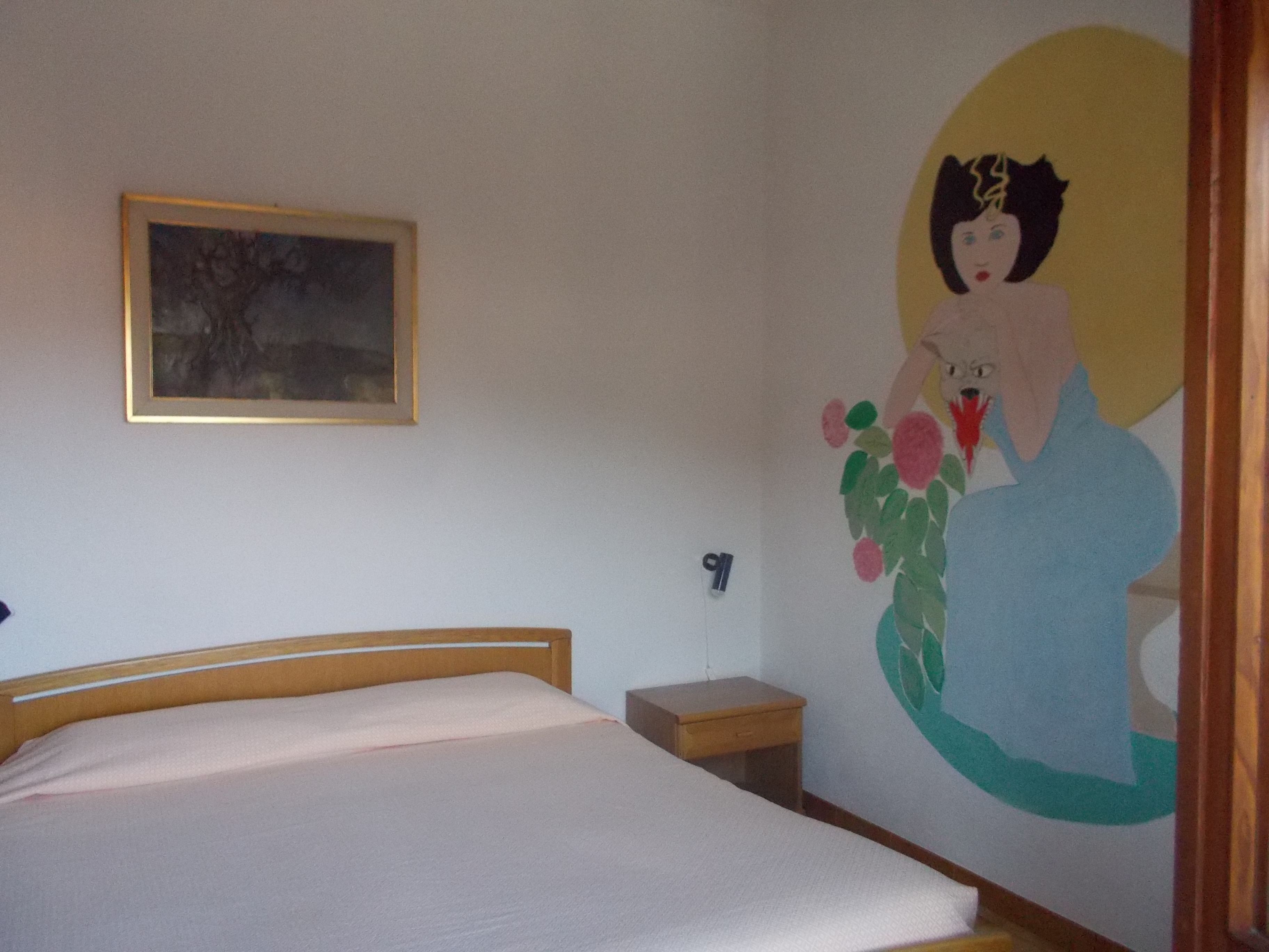 A bedroom with a bed, a small bedside table, a wall-mounted light switch, and a painting.