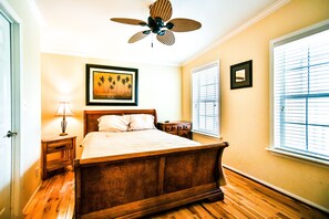 A bedroom with a wooden bed, a ceiling fan, a nightstand with a lamp, a framed picture on the wall, and a window with blinds. - Siesta Key Beach Home (Siesta Key)