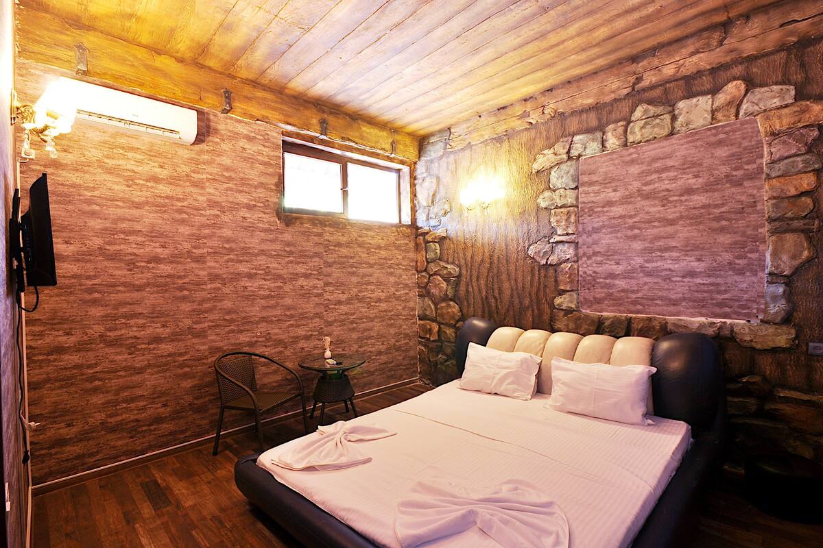 A bedroom with a wooden ceiling, stone walls, a bed with white linens, a small table with a chair, and a mounted TV.