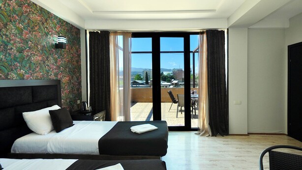 A hotel room with two beds, a balcony with a view of buildings and mountains, and a large window with curtains. - Marsel Hotel (Tbilisi)