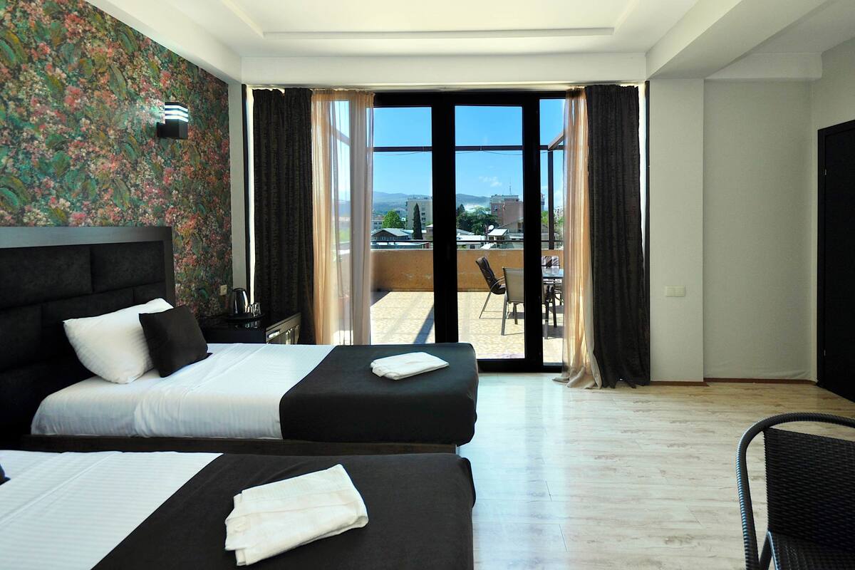 A hotel room with two beds, a balcony with a view of buildings and mountains, and a large window with curtains.