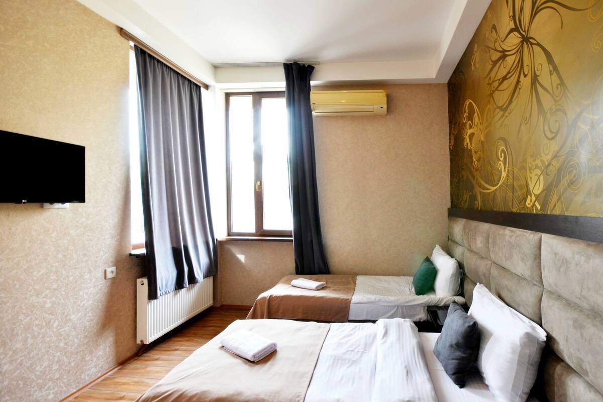 A hotel room with two beds, a flat-screen TV, a radiator, and a window with curtains.