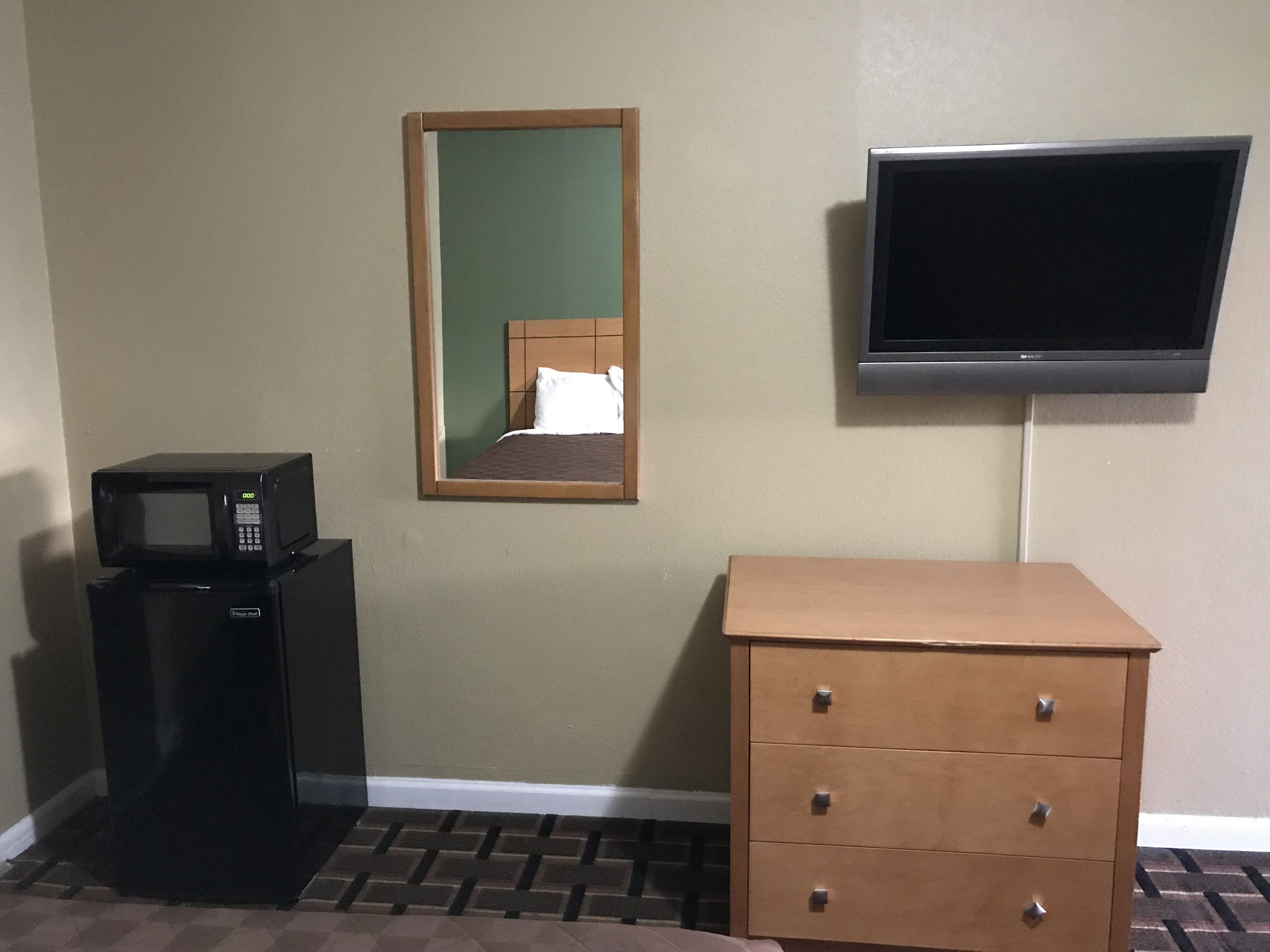 Habitación de hotel con microondas, pequeño refrigerador, cómoda de madera, espejo y televisor montado en la pared.