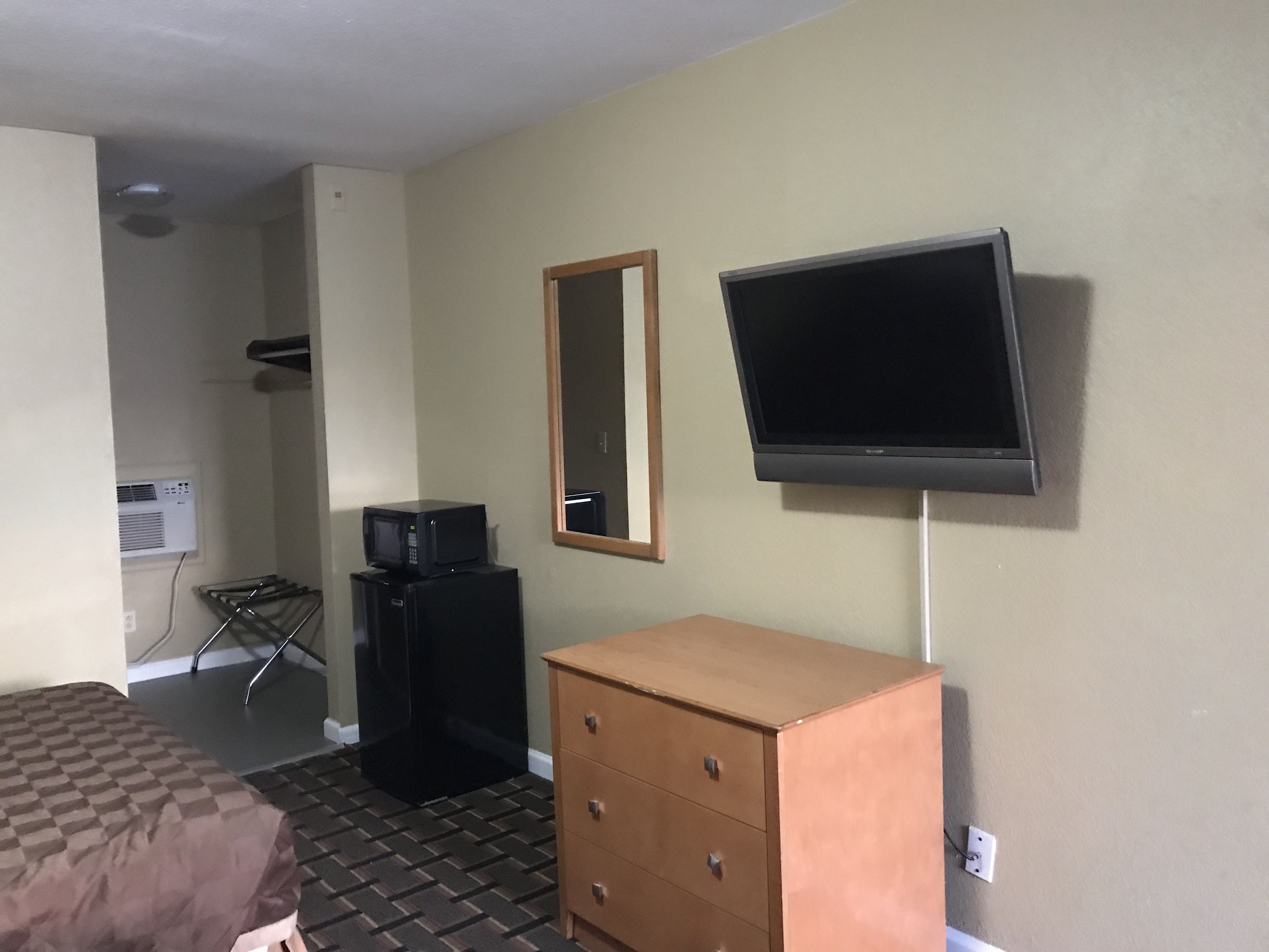 Una habitación de hotel con cama, cómoda, televisor montado en la pared y microondas sobre un mueble.