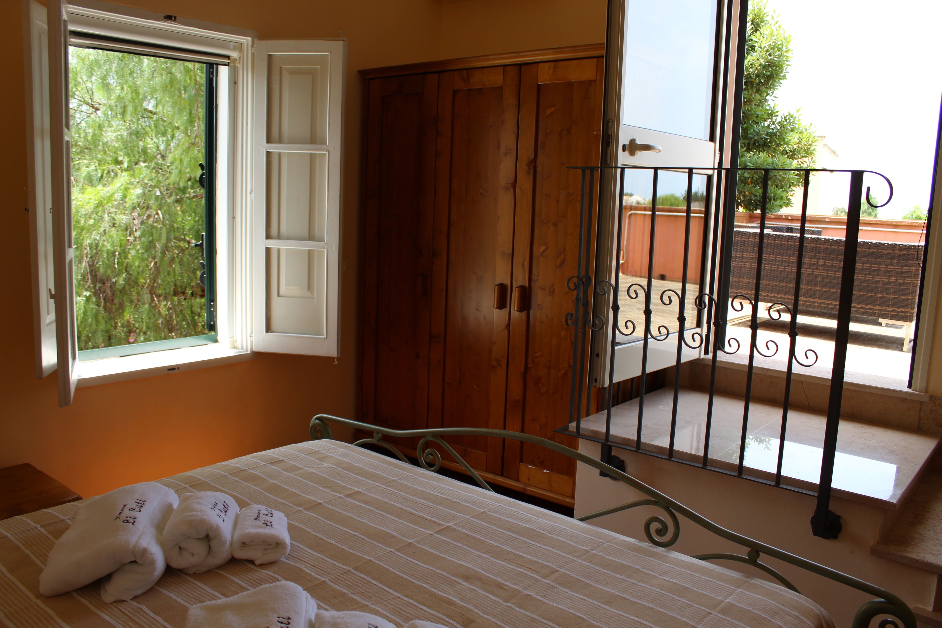 A bedroom with a bed, a window, a wooden wardrobe, and a balcony with a metal railing.