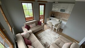 Una sala de estar moderna con un sofá seccional color beige, una alfombra pequeña y una zona de comedor con una mesa y sillas blancas. Se ve una cocina con gabinetes blancos y una ventana al fondo.