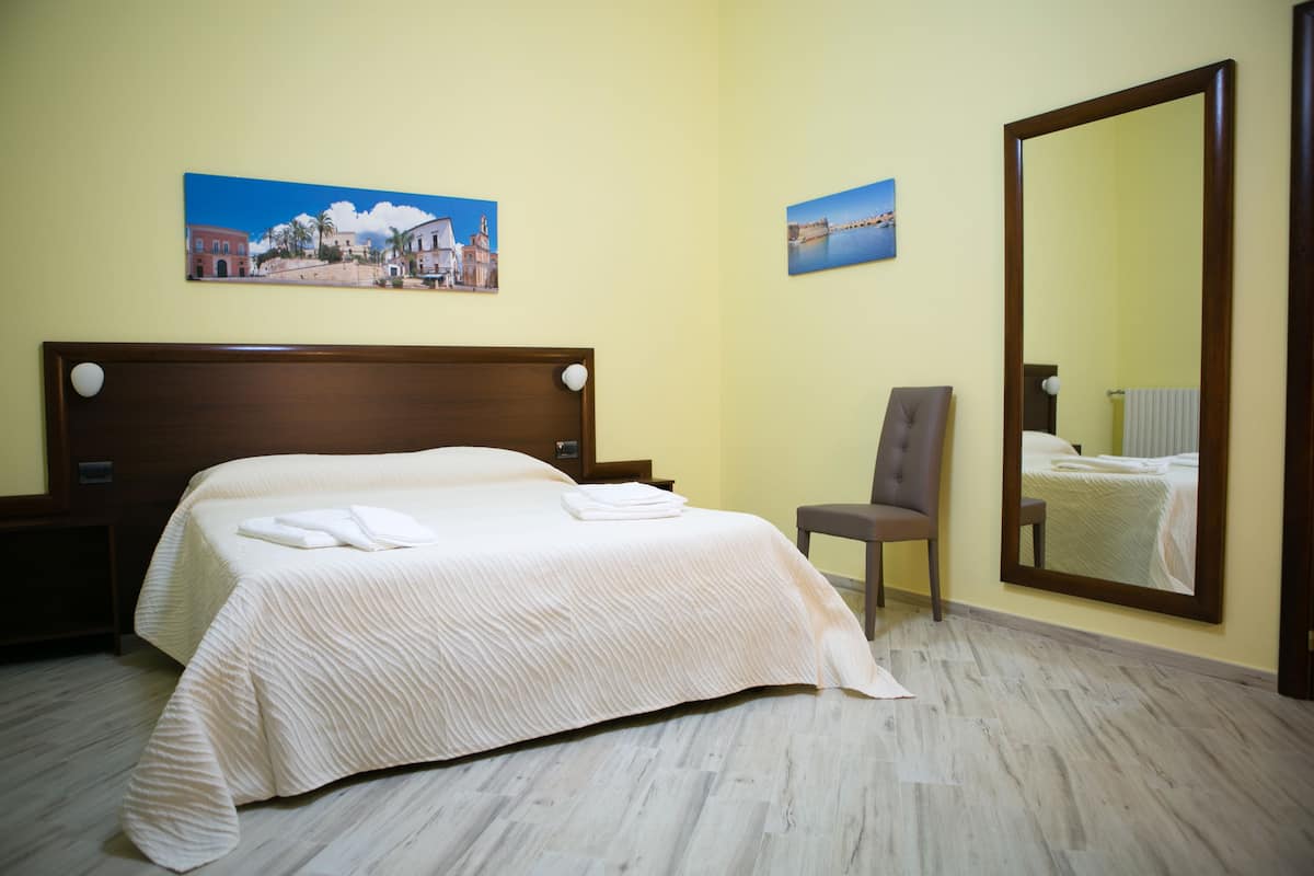 Quarto de hotel com uma cama grande, uma cadeira, uma mesa de cabeceira e duas pinturas penduradas na parede.