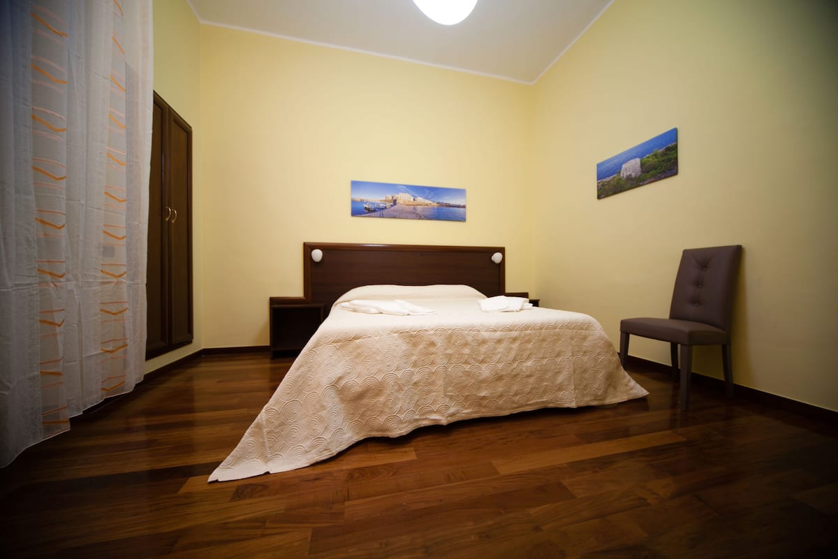 Um quarto com piso de madeira, uma cama com cabeceira, uma cadeira e duas pinturas na parede.