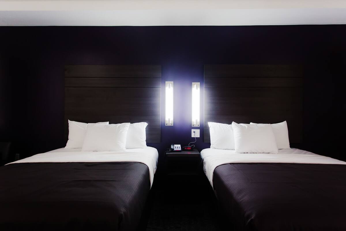 Habitación de hotel con dos camas, un cabecero oscuro y una mesita de noche con un reloj y un teléfono.