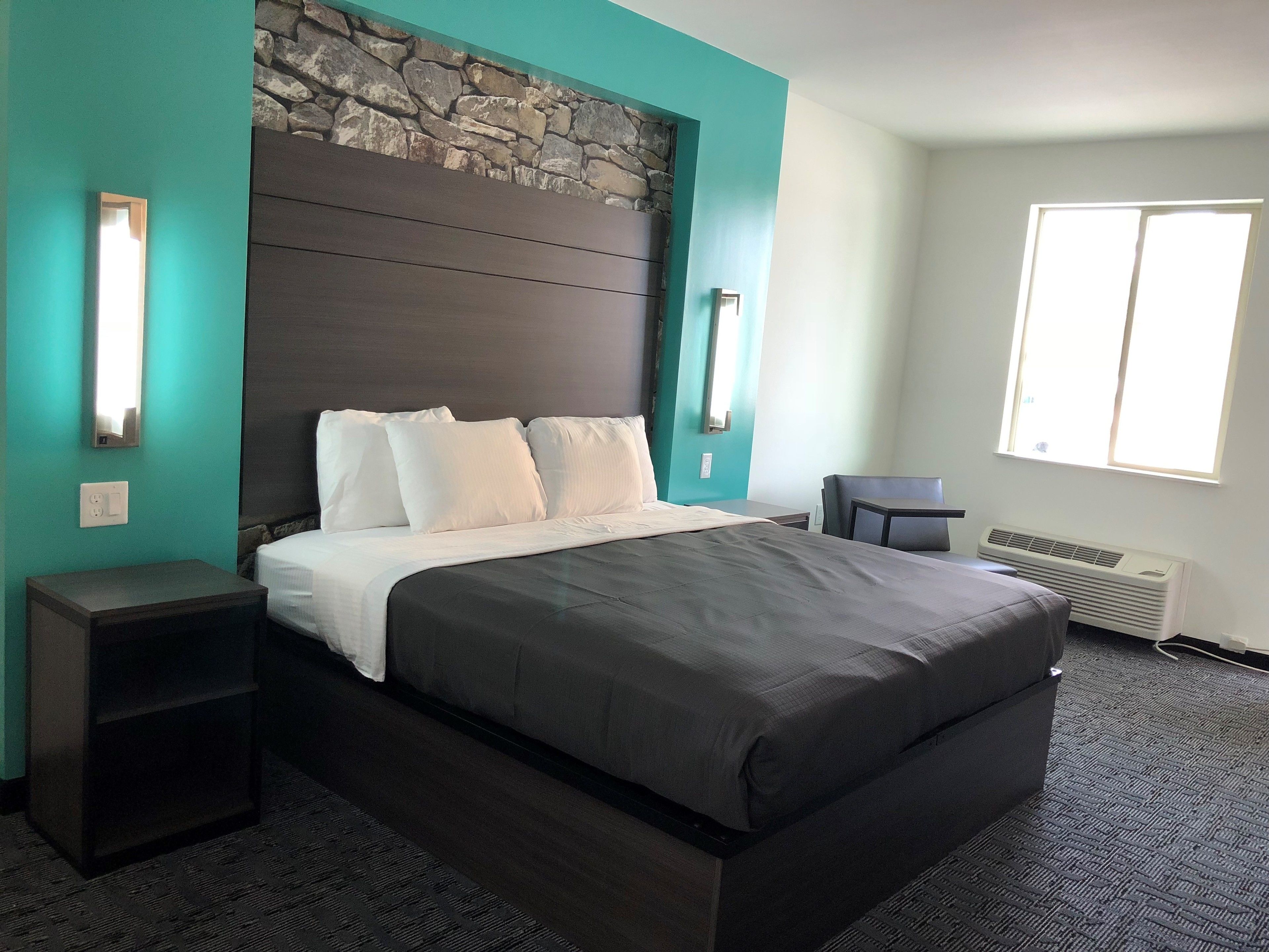 A hotel room with a bed, a nightstand, a chair, a window, and a stone wall.