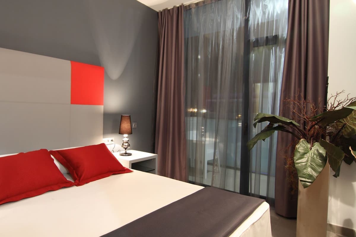 A hotel room with a large bed, red pillows, a bedside table with a lamp, a window with curtains, and a potted plant.