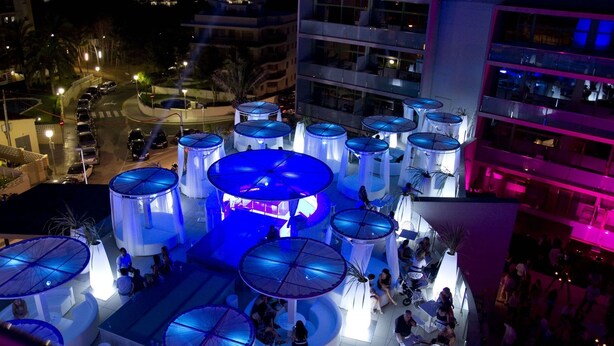 A rooftop bar with multiple blue umbrellas, a central bar area, and people socializing. - Aparthotel Four Elements Suites (Salou)