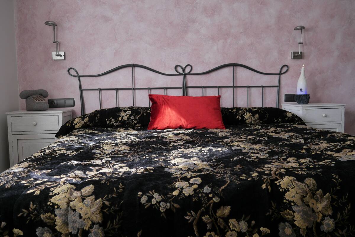 A bed with a black and gold floral bedspread, a red pillow, and a black metal headboard. There are two bedside tables with lamps and a telephone on one.