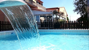 Une piscine agrémentée d’une cascade, entourée d’une clôture en bois et bordée d’un bâtiment doté de balcons.