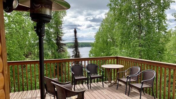 Eine Holzterrasse mit Korbstühlen und einem Tisch, mit Blick auf einen See und einen Wald.