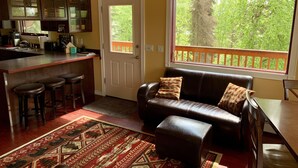 Ein gemütliches Wohnzimmer mit einem braunen Ledersofa, dazu passend einem Hocker und einem Teppich mit Muster. Links ist eine Küchenzeile mit Bar und Barhocker erkennbar.
