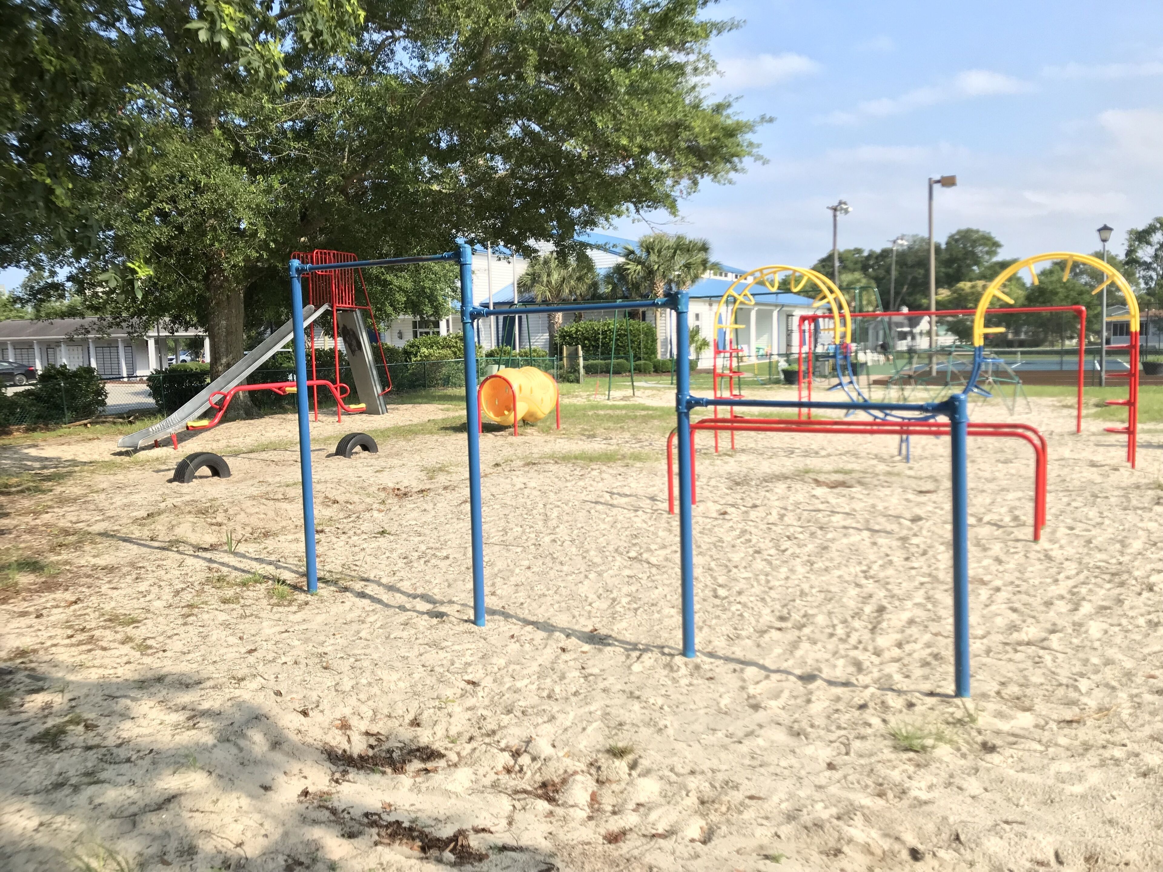 A playground with various play structures, including a slide and climbing frames, set in a sandy area with trees and a building in the background.