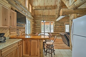Interior de una cabaña de troncos con una mesa de comedor de madera, sillas, un sofá y un refrigerador.