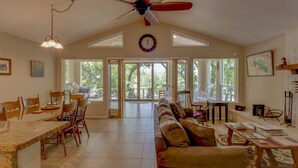 A spacious living room with a large ceiling fan, a dining area with wooden chairs and a granite table, and large windows offering a view of the outdoors.