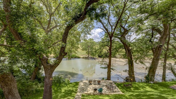 A serene outdoor seating area with stone steps, tables, and chairs overlooking a calm body of water, surrounded by lush trees.