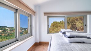 Ein Schlafzimmer mit einem großen Bett, zwei Fenstern mit Jalousien und Blick auf eine Landschaft.