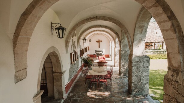 A corridor with stone arches, a central table with chairs, and a cross on the wall. - Meson Real de la Plata (El Marques)