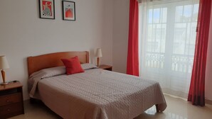 A neatly made bed with a red pillow, two bedside tables with lamps, and a window with red curtains.