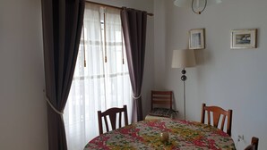 A dining area with a round table covered in a floral tablecloth, two wooden chairs, a lamp, and a window with curtains.