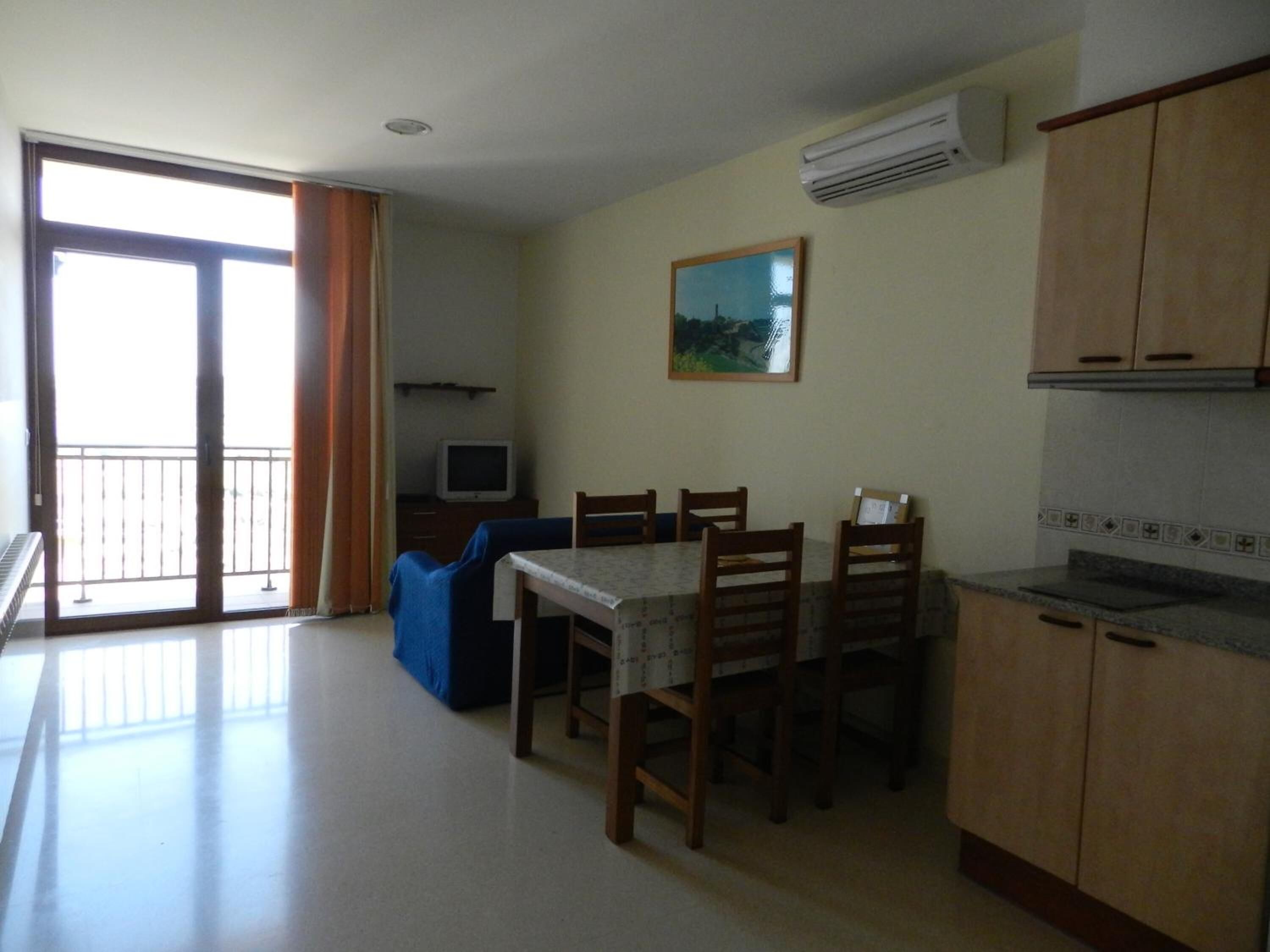 A small, bright kitchen with wooden cabinets, a dining table with chairs, a TV, and a large window with curtains.