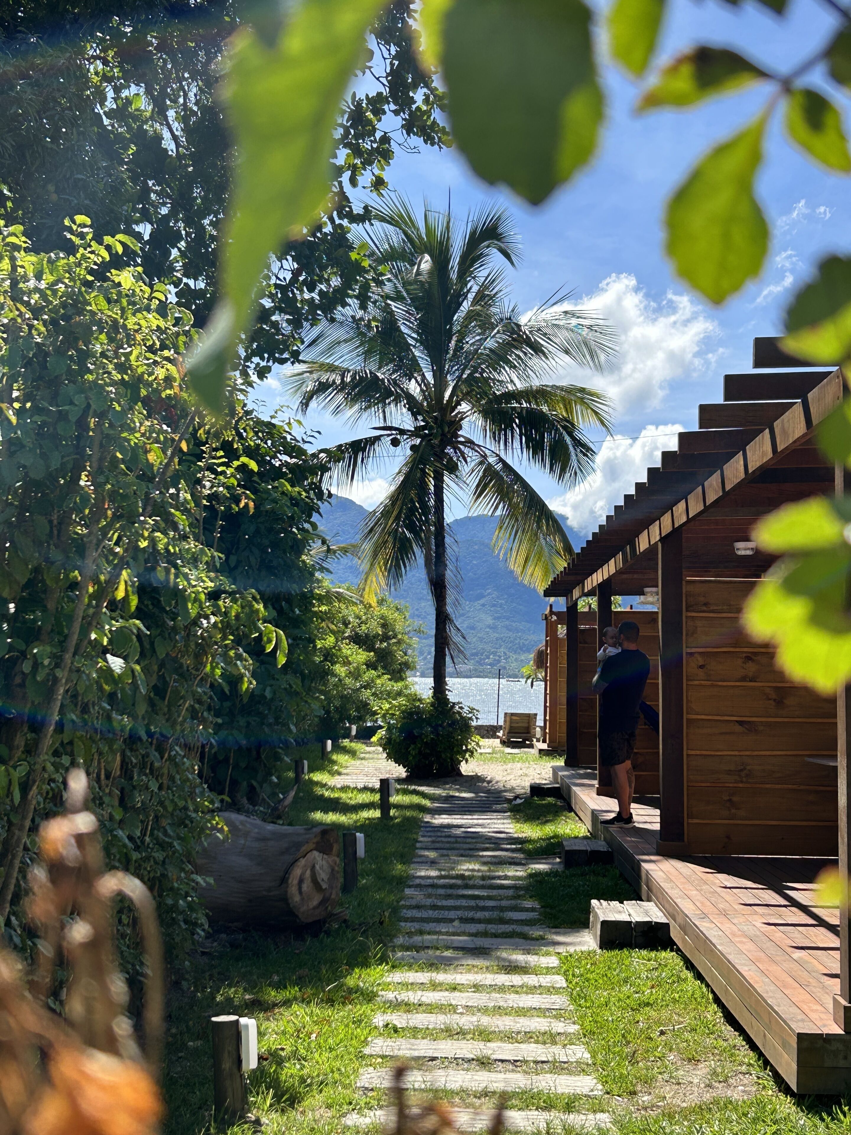 Ein Holzweg führt zu einem überdachten Außenbereich mit einer Palme und Bergen im Hintergrund.