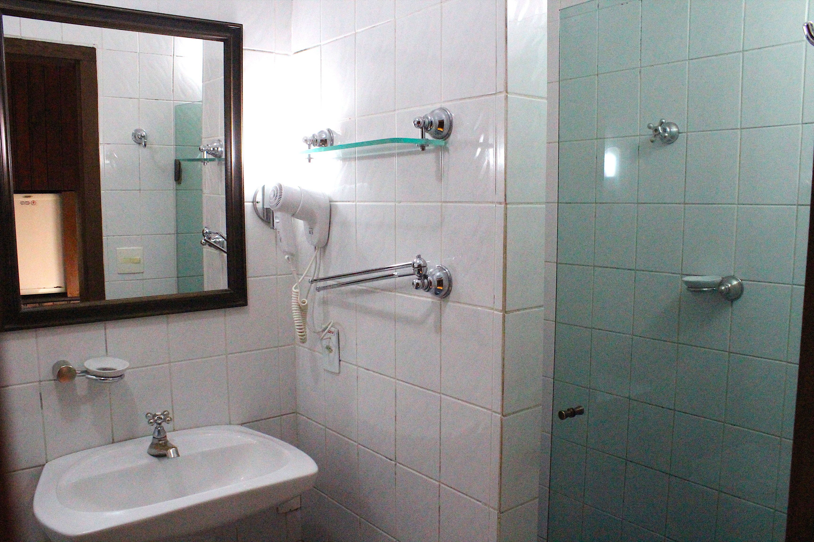 A bathroom with a white sink, a mirror, a hairdryer, and a shower with a glass shelf.